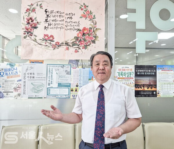 ▴인터뷰를 마치고 최귀옥 원장이 ‘도∙봉∙문∙화∙원’ 오행시 캘리그라피 앞에서 사진을 찍고 있는 모습(오행시 뜻: (도)는 길이요 변하지 않는 진리, (봉)은 하늘을 받치는 기둥, (문)은 배움의 씨앗 뿌리고 드나드는 곳, (화)는 결실을 위한 삶의 무늬요 여유, (원)은 따로 또 같이 일구어낸 여유와 희망)